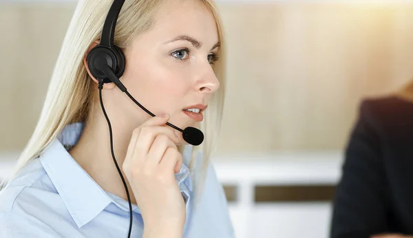 Blond affärskvinna sitter och kommuniceras med headset i soligt call center kontor — Stockfoto