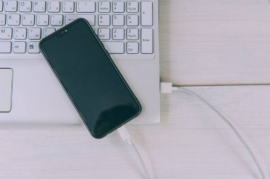 Akıllı telefon beyaz, ahşap bir masada dizüstü bilgisayardan şarj oluyor.