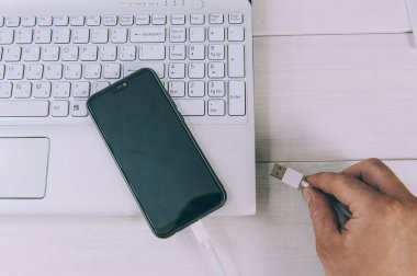 Adam akıllı telefonu beyaz bir masada dizüstü bilgisayarla şarj ediyor.