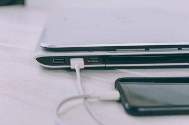 Akıllı telefon beyaz, ahşap bir masada dizüstü bilgisayardan şarj oluyor.