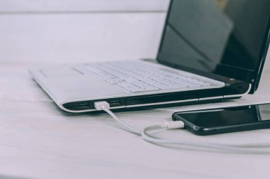 Akıllı telefon beyaz, ahşap bir masada dizüstü bilgisayardan şarj oluyor.