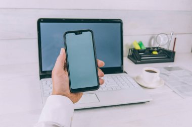 Model Teknolojisi. İşadamı elinde bir akıllı telefon tutuyor, dizüstü bilgisayarın arka planında ve ahşap bir masada kahve.