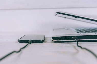 Akıllı telefon beyaz, ahşap bir masada dizüstü bilgisayardan şarj oluyor.