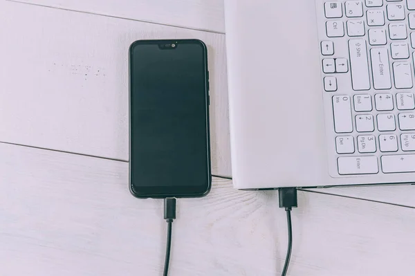 Akıllı telefon beyaz, ahşap bir masada dizüstü bilgisayardan şarj oluyor.