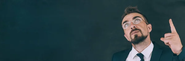 Gömlekli ve kravatlı bir iş adamı, koyu bir arka planda, gözlüklü, en üstte bir parmağını gösteriyor.