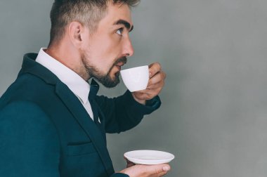 Ceketli iş adamı içer ve elinde gri arka planda kahve olan bir fincan tutar.