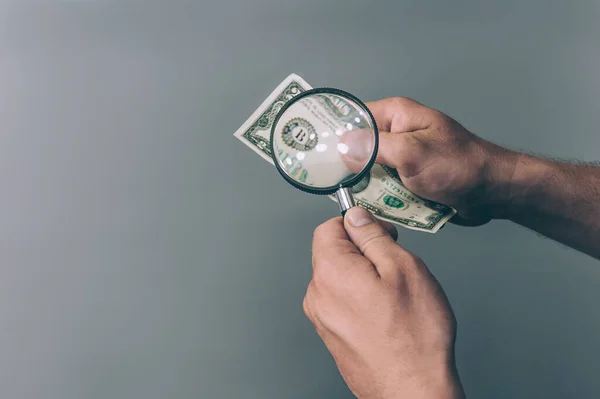 Bir adam elinde bir büyüteç tutuyor ve gri arka planda sahte para var mı diye dolarları kontrol ediyor.