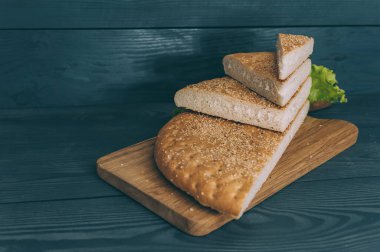 Gri bir masanın üzerindeki kesme tahtasına dilimlenmiş yeşillerle bir somun ekmek.