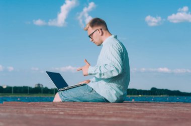 Gözlüklü ve gömlekli bir adam temiz havada dizüstü bilgisayarda çalışıyor, nehrin arka planına karşı.