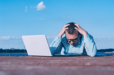 Gözlüklü ve gömlekli bir adam temiz havada dizüstü bilgisayarda çalışıyor, nehrin arka planına karşı.