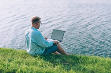 Gözlüklü ve gömlekli bir adam temiz havada dizüstü bilgisayarda çalışıyor, nehrin arka planına karşı.