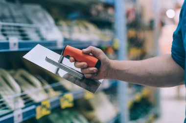 Hırdavatçıdaki alıcı malları seçer. Adamın biri elinde metal bir spatula tutuyor.