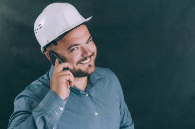 İnşaat kasklı bir adam telefonda koyu bir arkaplanda konuşuyor.