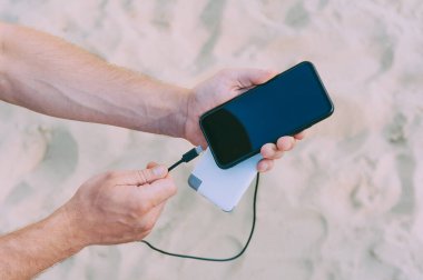 Kumsalın arka planındaki bir adamın elinde taşınabilir bir şarj aleti. Powerbank telefonu şarj ediyor