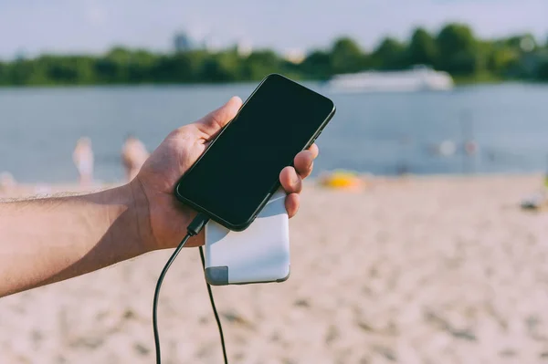 Portable charger in the hands of a man on the background of the beach. Powerbank charges the phone