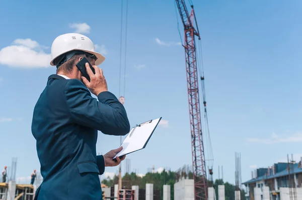 Ceketli bir şef ve ellerinde belgelerle bir kule vincinin arka planına ve bulutlu gökyüzüne bakan miğfer akıllı bir telefonla konuşuyor.