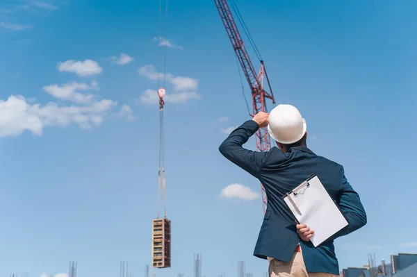 Ceketli bir şef ve ellerinde belgeler olan bir miğfer kule vincinin arka planına ve bulutlu gökyüzüne karşı. Ev inşa denetimi