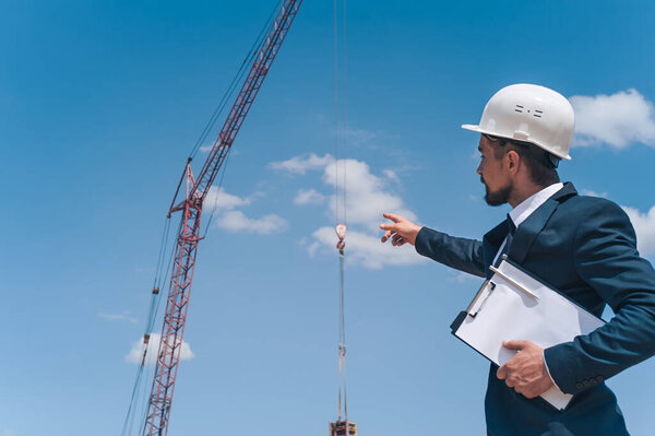 Начальник в куртке и строительном шлеме с документами в руках, на фоне башенного крана и неба с облаками. Проверка строительства домов