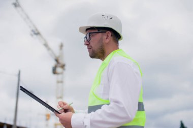 Yelek giymiş bir şef ve ellerinde belgeler olan bir miğfer kule vincinin arka planına ve bulutlu gökyüzüne karşı. Ev inşa denetimi