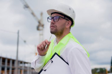 Yelek giymiş bir şef ve ellerinde belgeler olan bir miğfer kule vincinin arka planına ve bulutlu gökyüzüne karşı. Ev inşa denetimi