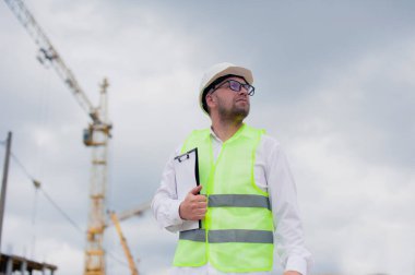Yelek giymiş bir şef ve ellerinde belgeler olan bir miğfer kule vincinin arka planına ve bulutlu gökyüzüne karşı. Ev inşa denetimi