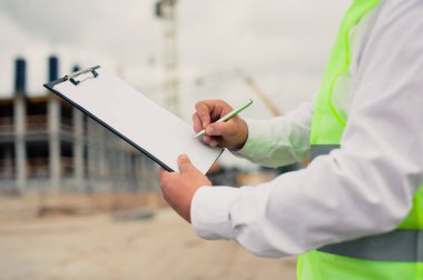 Yelek giymiş bir şef ve ellerinde belgeler olan bir miğfer kule vincinin arka planına ve bulutlu gökyüzüne karşı. Ev inşa denetimi