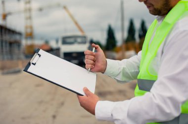 Yelek giymiş bir şef ve ellerinde belgeler olan bir miğfer kule vincinin arka planına ve bulutlu gökyüzüne karşı. Ev inşa denetimi