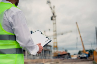 Yelek giymiş bir şef ve ellerinde belgeler olan bir miğfer kule vincinin arka planına ve bulutlu gökyüzüne karşı. Ev inşa denetimi