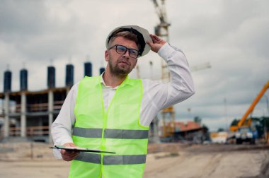 Yelek giymiş bir şef ve ellerinde belgeler olan bir miğfer kule vincinin arka planına ve bulutlu gökyüzüne karşı. Ev inşa denetimi