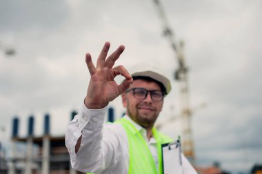 Yelek giymiş bir şef ve ellerinde belgeler olan bir miğfer kule vincinin arka planına ve bulutlu gökyüzüne karşı. Ev inşa denetimi