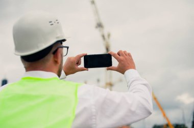 Ceketli bir şef ve ellerinde akıllı telefon olan bir miğfer kule vincinin arka planına ve bulutlu gökyüzüne karşı.