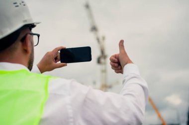 Ceketli bir şef ve ellerinde akıllı telefon olan bir miğfer kule vincinin arka planına ve bulutlu gökyüzüne karşı.