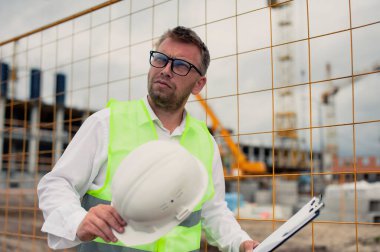 Yelek giymiş bir şef ve ellerinde belgeler olan bir miğfer kule vincinin arka planına ve bulutlu gökyüzüne karşı. Ev inşa denetimi