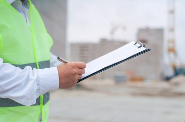 Yelek giymiş bir şef ve ellerinde belgeler olan bir miğfer kule vincinin arka planına ve bulutlu gökyüzüne karşı. Ev inşa denetimi