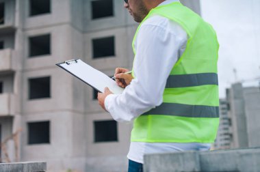 Yelek giymiş bir şef ve ellerinde belgeler olan bir miğfer kule vincinin arka planına ve bulutlu gökyüzüne karşı. Ev inşa denetimi