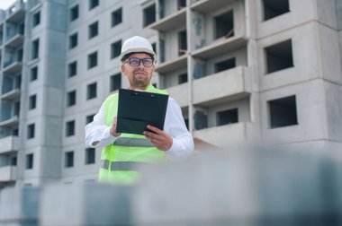 Yelek giymiş bir şef ve ellerinde belgeler olan bir miğfer kule vincinin arka planına ve bulutlu gökyüzüne karşı. Ev inşa denetimi
