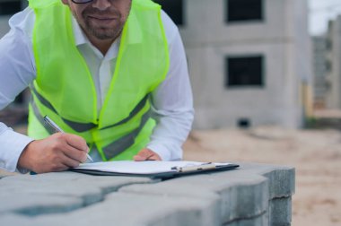 Yelek giymiş bir şef ve ellerinde belgeler olan bir miğfer kule vincinin arka planına ve bulutlu gökyüzüne karşı. Ev inşa denetimi
