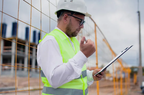 Yelek giymiş bir şef ve ellerinde belgeler olan bir miğfer kule vincinin arka planına ve bulutlu gökyüzüne karşı. Ev inşa denetimi