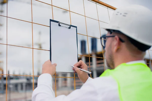 Yelek giymiş bir şef ve ellerinde belgeler olan bir miğfer kule vincinin arka planına ve bulutlu gökyüzüne karşı. Ev inşa denetimi