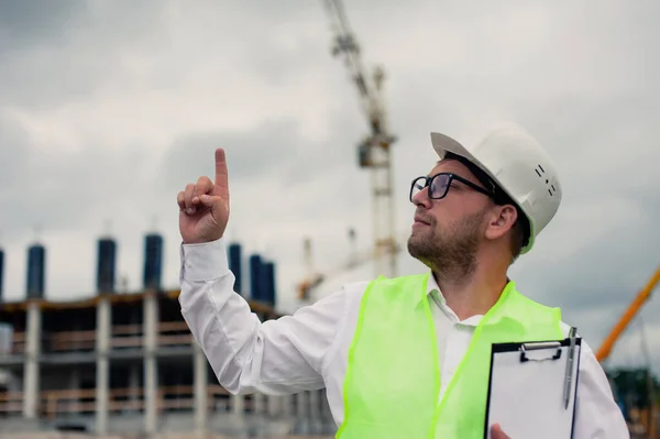 Yelek giymiş bir şef ve ellerinde belgeler olan bir miğfer kule vincinin arka planına ve bulutlu gökyüzüne karşı. Ev inşa denetimi
