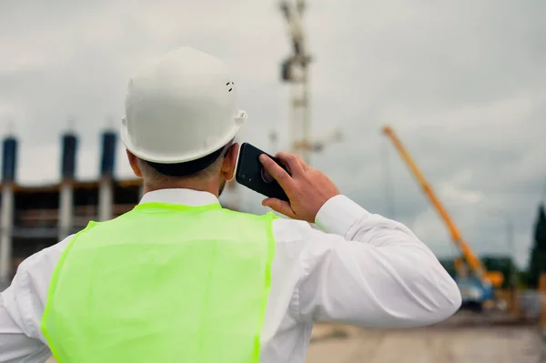 Ceketli bir şef ve ellerinde akıllı telefon olan bir miğfer kule vincinin arka planına ve bulutlu gökyüzüne karşı.