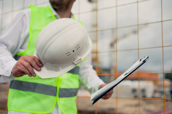 Начальник в жилете и строительном шлеме с документами в руках, на фоне башенного крана и неба с облаками. Проверка строительства домов