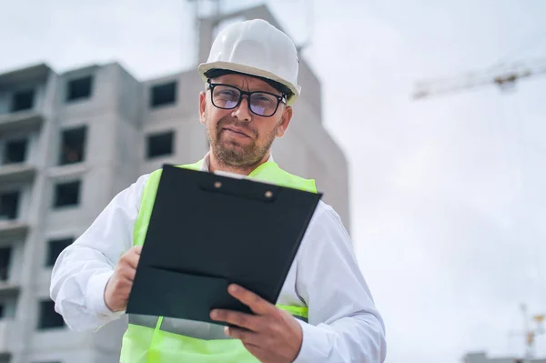 Yelek giymiş bir şef ve ellerinde belgeler olan bir miğfer kule vincinin arka planına ve bulutlu gökyüzüne karşı. Ev inşa denetimi