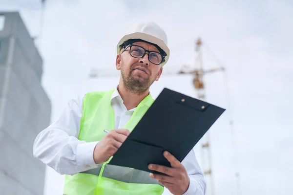 Yelek giymiş bir şef ve ellerinde belgeler olan bir miğfer kule vincinin arka planına ve bulutlu gökyüzüne karşı. Ev inşa denetimi
