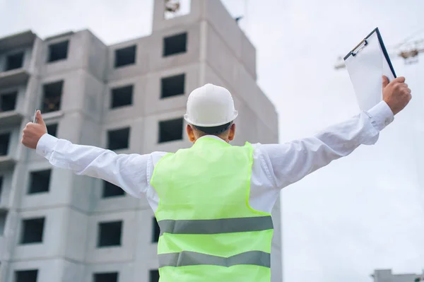 Chief in a vest and a construction helmet with documents in his hands, against the background of a tower crane and the sky with clouds. House construction inspection