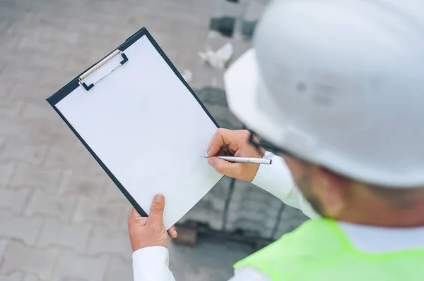 Yelek giymiş bir şef ve ellerinde belgeler olan bir miğfer kule vincinin arka planına ve bulutlu gökyüzüne karşı. Ev inşa denetimi