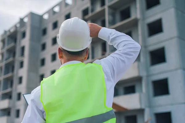 Chief in a vest and a construction helmet with documents in his hands, against the background of a tower crane and the sky with clouds. House construction inspection