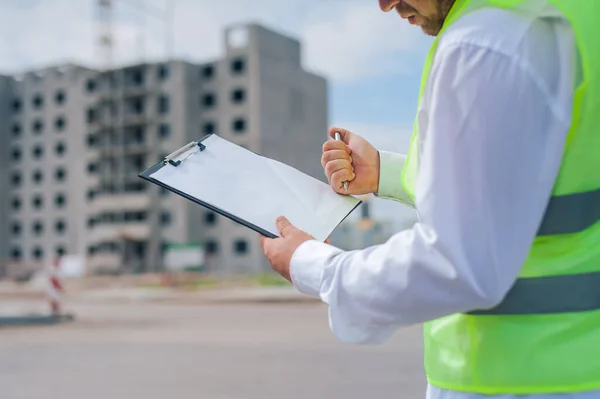 Yelek giymiş bir şef ve ellerinde belgeler olan bir miğfer kule vincinin arka planına ve bulutlu gökyüzüne karşı. Ev inşa denetimi