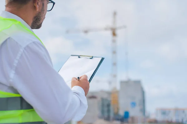 Yelek giymiş bir şef ve ellerinde belgeler olan bir miğfer kule vincinin arka planına ve bulutlu gökyüzüne karşı. Ev inşa denetimi