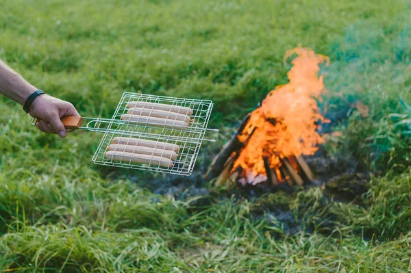 Człowiek Kratą Ręku Przygotowuje Kiełbaski Ogniu Piknik Przyrodzie — Zdjęcie stockowe